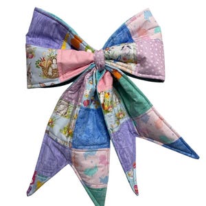 May include: A large, handmade fabric bow in a patchwork design. The bow features various pastel colors, including pink, blue, and lavender, with Easter-themed patterns. The bow is likely used for decoration or as a hair accessory.