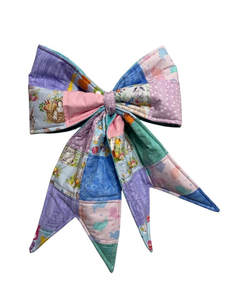 May include: A large, handmade fabric bow in a patchwork design. The bow features various pastel colors, including pink, blue, and lavender, with Easter-themed patterns. The bow is likely used for decoration or as a hair accessory.