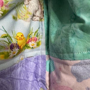 May include: A patchwork fabric with Easter-themed patterns. The fabric features a light blue section with a bunny and chick, a purple section, a pink section with a bunny, and a green section. The fabric is likely for crafts or sewing projects.