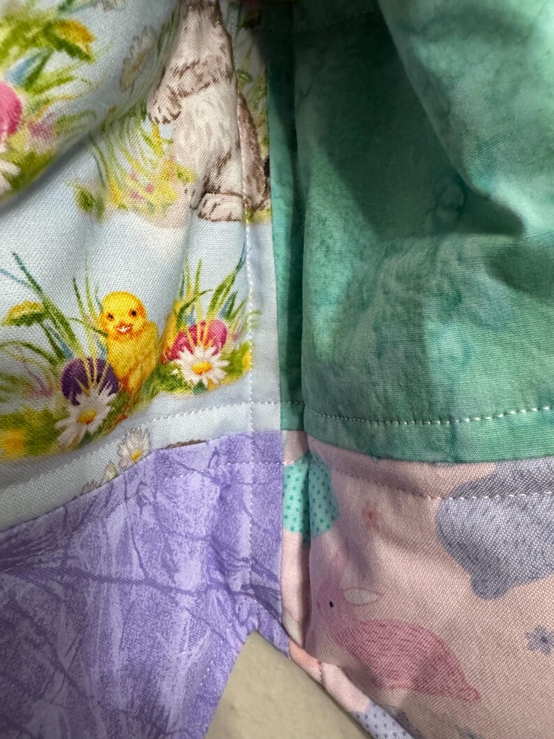 May include: A patchwork fabric with Easter-themed patterns. The fabric features a light blue section with a bunny and chick, a purple section, a pink section with a bunny, and a green section. The fabric is likely for crafts or sewing projects.