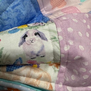 May include: Close-up of a patchwork fabric with various patterns. The fabric features a blue, pink, and purple color scheme. One section has a bunny print, another has polka dots, and another has a bunny illustration. The fabric is quilted.