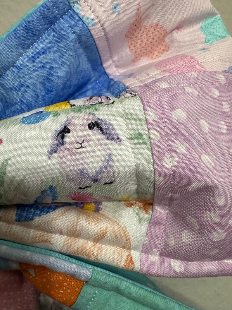 May include: Close-up of a patchwork fabric with various patterns. The fabric features a blue, pink, and purple color scheme. One section has a bunny print, another has polka dots, and another has a bunny illustration. The fabric is quilted.