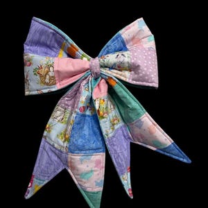 May include: A large, colorful bow made of patchwork fabric. The bow features various pastel colors and patterns, including bunnies, eggs, and floral designs. The bow is likely handmade and has a decorative appearance.