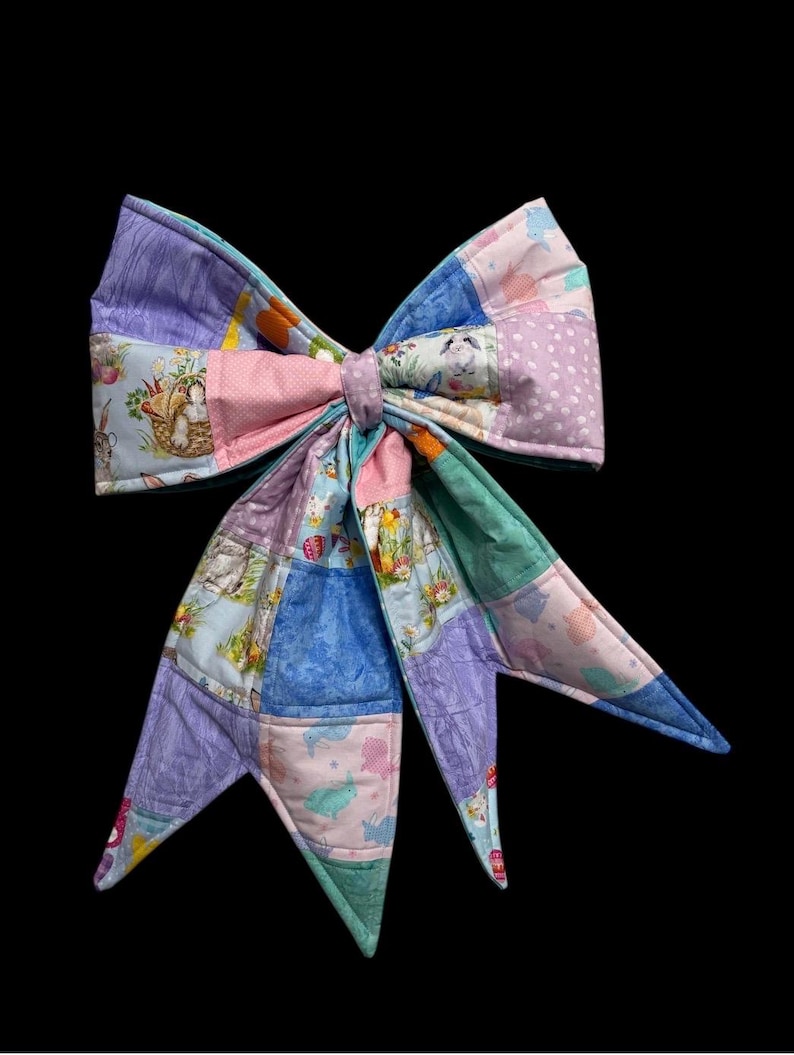 May include: A large, colorful bow made of patchwork fabric. The bow features various pastel colors and patterns, including bunnies, eggs, and floral designs. The bow is likely handmade and has a decorative appearance.