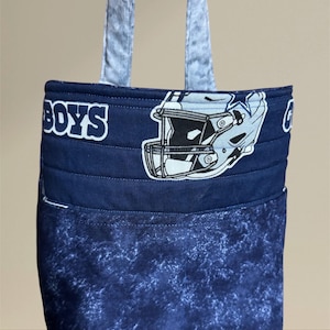 May include: A navy blue tote bag with a light blue handle. The bag features a football helmet graphic and the word "BOYS" in white. The lower portion of the bag has a blue and white marbled pattern.