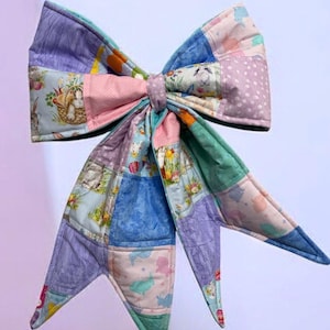 May include: A large, quilted bow in pastel colors. The bow is made of patchwork squares featuring bunnies, flowers, and polka dots. The bow's center is a solid pink, and the tails are pointed.