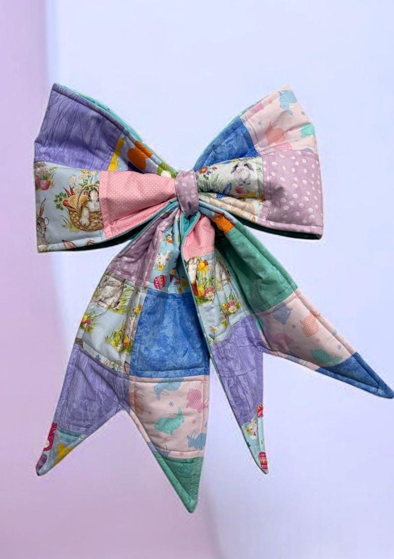 May include: A large, quilted bow in pastel colors. The bow is made of patchwork squares featuring bunnies, flowers, and polka dots. The bow's center is a solid pink, and the tails are pointed.