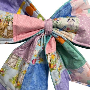 May include: A large, quilted bow in pastel colors. The bow features a patchwork design with various patterns, including bunnies, flowers, and polka dots. The bow is tied in the center with a matching fabric band. The overall theme is Easter or spring.
