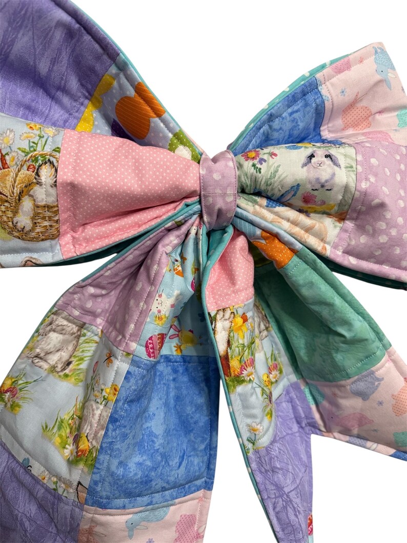May include: A large, quilted bow in pastel colors. The bow features a patchwork design with various patterns, including bunnies, flowers, and polka dots. The bow is tied in the center with a matching fabric band. The overall theme is Easter or spring.