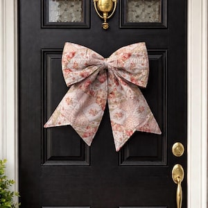 May include: A large, decorative bow made of floral patterned fabric, in shades of pink, cream, and red, is attached to a black front door. The bow is the focal point, with a brass door knocker and handle adding accents. This festive bow is perfect for seasonal home decor.