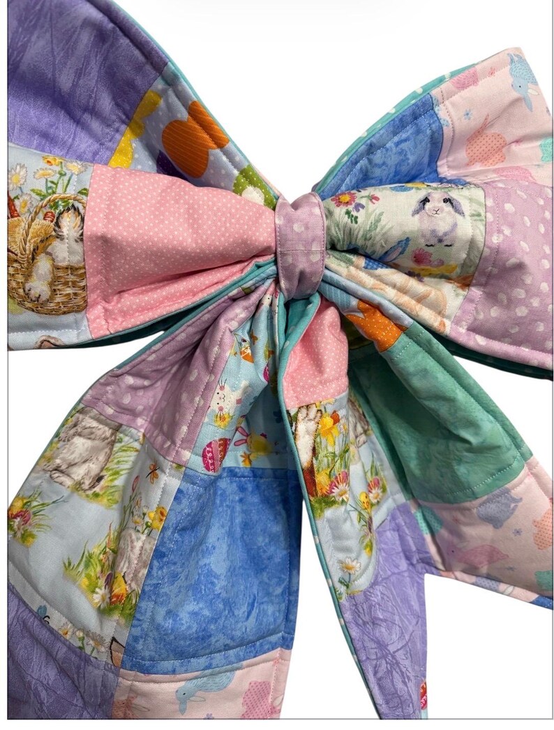 May include: A large, colorful fabric bow featuring a patchwork design. The bow incorporates various pastel colors, including pink, blue, purple, and green. The fabric is decorated with Easter-themed patterns, such as bunnies and floral motifs, creating a festive and decorative item.