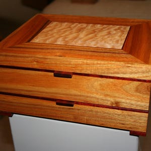 May include: A rectangular wooden box featuring two drawers with dark brown horizontal accents. The top surface displays a lighter, wavy wood grain pattern framed by a darker, reddish-brown border. The box has a smooth, polished finish.