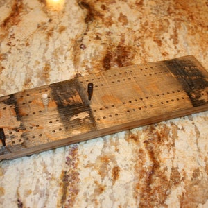 May include: A wooden cribbage board with a dark brown stain and multiple holes for pegs. The board has four pegs on it, two white and two brown.