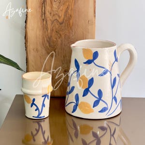 Może przedstawiać: Kremowy ceramiczny dzbanek i pasująca filiżanka, oba zdobione wzorem żółtych cytryn i niebieskich liści. Dzbanek ma uchwyt i dzióbek. Filiżanka jest cylindryczna. W tle widoczna jest drewniana deska do krojenia.