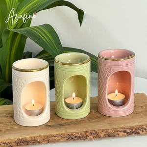 Op de afbeelding: Drie keramische kaarshouders in crème, groen en roze, elk met een gouden rand en een brandende waxinelichtkaars erin. De houders hebben decoratieve patronen en worden op een houten oppervlak weergegeven. Het woord "Azafine" is cursief geschreven.