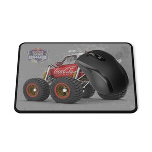May include: A red, white and black Red Bull Sand Scramble monster truck with a Coca-Cola logo on the side. A black computer mouse is on top of the truck. The background is a grey, textured surface.