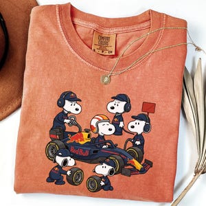 May include: A terracotta-colored t-shirt featuring a graphic of Snoopy and friends as a Red Bull Formula 1 pit crew. The shirt has a Comfort Colors brand label in the neck, showing size S. The graphic includes a race car and the Red Bull logo.