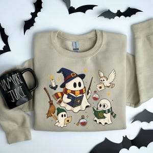 May include: A sand-colored crewneck sweatshirt featuring a graphic of three ghosts dressed as wizards, holding books, a wand, and potions, with an owl and broomstick. Beside it is a black mug with the white text "VAMPIRE JUICE" printed on it.