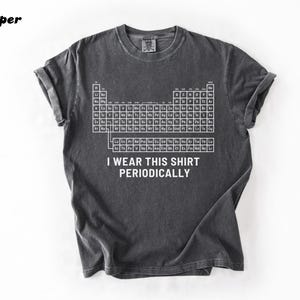 May include: Dark gray t-shirt featuring a white periodic table design. The shirt includes the text "I wear this shirt periodically." The shirt has rolled-up sleeves and a crew neck.