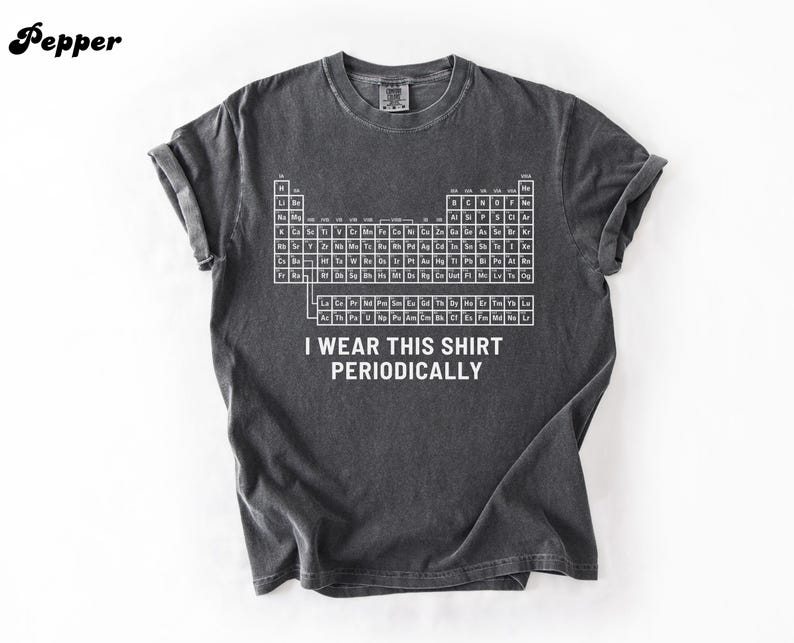 May include: Dark gray t-shirt featuring a white periodic table design. The shirt includes the text "I wear this shirt periodically." The shirt has rolled-up sleeves and a crew neck.