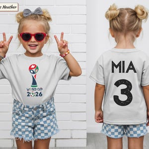 May include: An athletic heather gray t-shirt featuring a World Cup 2026 graphic on the front with a soccer ball icon. The back of the shirt displays the name MIA in black capital letters above the large black number 3 with a distressed texture.