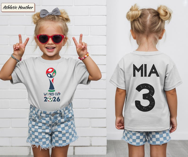 May include: An athletic heather gray t-shirt featuring a World Cup 2026 graphic on the front with a soccer ball icon. The back of the shirt displays the name MIA in black capital letters above the large black number 3 with a distressed texture.
