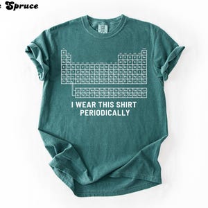 May include: Teal-colored t-shirt featuring a white periodic table design. The shirt includes the text "I WEAR THIS SHIRT PERIODICALLY" in white. The shirt is made of a soft material and has short sleeves.