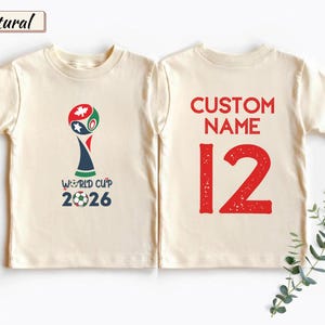 May include: A natural-colored t-shirt shown from the front and back. The front features a World Cup 2026 graphic with a soccer ball. The back displays the red text CUSTOM NAME above the large red number 12 in a distressed font style.
