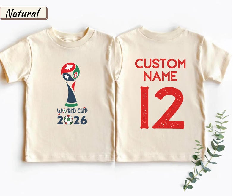 May include: A natural-colored t-shirt shown from the front and back. The front features a World Cup 2026 graphic with a soccer ball. The back displays the red text CUSTOM NAME above the large red number 12 in a distressed font style.