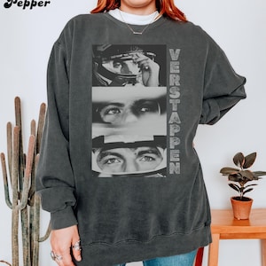 May include: Dark gray sweatshirt featuring a black and white image of a race car driver's face in three sections. The word "VERSTAPPEN" is printed vertically on the right side of the sweatshirt. The brand name "Pepper" is printed in the upper left corner.