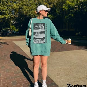 May include: A teal-colored sweatshirt dress with the word "VERSTAPPEN" printed vertically on the front, along with a graphic of a person's eyes. The image also shows a white baseball cap, sunglasses, and white sneakers.