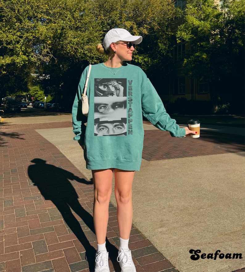 May include: A teal-colored sweatshirt dress with the word "VERSTAPPEN" printed vertically on the front, along with a graphic of a person's eyes. The image also shows a white baseball cap, sunglasses, and white sneakers.
