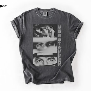 May include: Dark gray t-shirt featuring a black and white graphic of a person's eyes and face, with the name "VERSTAPPEN" printed vertically on the right side. The shirt has rolled-up sleeves and a crew neck.