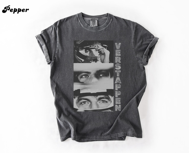May include: Dark gray t-shirt featuring a black and white graphic of a person's eyes and face, with the name "VERSTAPPEN" printed vertically on the right side. The shirt has rolled-up sleeves and a crew neck.