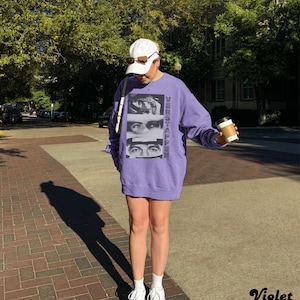 May include: A violet-colored sweatshirt with the word "VERSTAPPEN" printed vertically on the right side and a graphic of three sets of eyes. The person is wearing a white cap, sunglasses, and white sneakers. They are holding a coffee cup.
