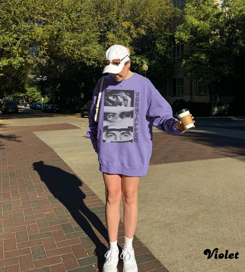 May include: A violet-colored sweatshirt with the word "VERSTAPPEN" printed vertically on the right side and a graphic of three sets of eyes. The person is wearing a white cap, sunglasses, and white sneakers. They are holding a coffee cup.