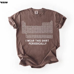 May include: Espresso-colored t-shirt with a white periodic table graphic and the text "I WEAR THIS SHIRT PERIODICALLY." The shirt has rolled sleeves and a relaxed fit. The shirt is made of a soft, comfortable material.