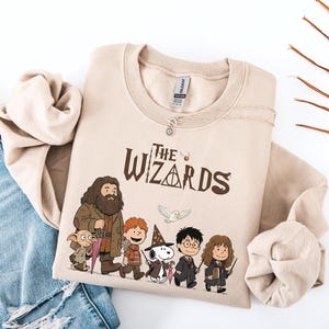 May include: A sand-colored crewneck sweatshirt featuring a graphic print with the text The Wizards and illustrations of characters including a large bearded figure, a small elf, a boy with red hair, a dog in a wizard hat, a boy with glasses, and a girl.