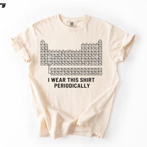 May include: Ivory-colored t-shirt featuring a black periodic table design. The shirt includes the text "I wear this shirt periodically" in a bold, sans-serif font. The shirt is made of a soft material and has short sleeves.