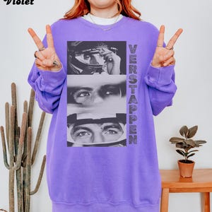 May include: A violet sweatshirt featuring a black and white image of a race car driver. The image is vertically stacked with the word "VERSTAPPEN" printed on the right side. The word "Violet" is printed in the upper left corner.