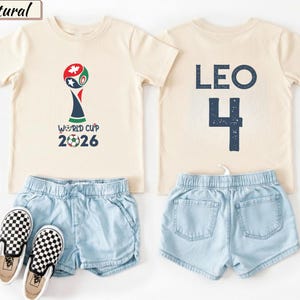 May include: Two natural-colored t-shirts, light blue denim shorts, and black and white checkered slip-on shoes. One shirt features a World Cup 2026 graphic with a soccer ball. The other shirt displays the name LEO and the number 4 in dark blue.