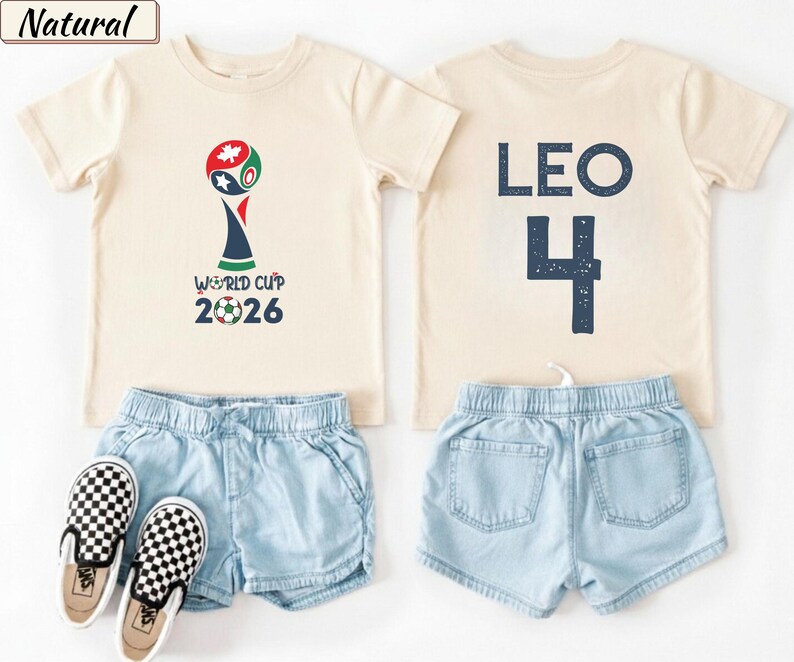 May include: Two natural-colored t-shirts, light blue denim shorts, and black and white checkered slip-on shoes. One shirt features a World Cup 2026 graphic with a soccer ball. The other shirt displays the name LEO and the number 4 in dark blue.