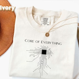 May include: Ivory-colored t-shirt with the text "CORE OF EVERYTHING" and a circuit board graphic. A gold necklace with a pendant rests on the shirt. The shirt is folded and displayed with a brown hat.