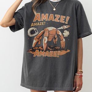 May include: Dark gray t-shirt with the word "AMAZE!" in orange, along with a cartoon illustration of a space crab, a rocket, and a space helmet. The shirt has a relaxed fit.