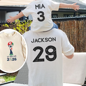 May include: A sand-natural colored t-shirt featuring the name JACKSON and the number 29 in black on the back. A smaller t-shirt in the same color displays the name MIA and number 3. A circular inset shows the World Cup 2026 logo in color.