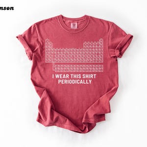 May include: Crimson-colored t-shirt featuring a white periodic table of elements graphic. The text "I wear this shirt periodically" is printed below the table. The shirt has rolled-up sleeves and a crew neck.
