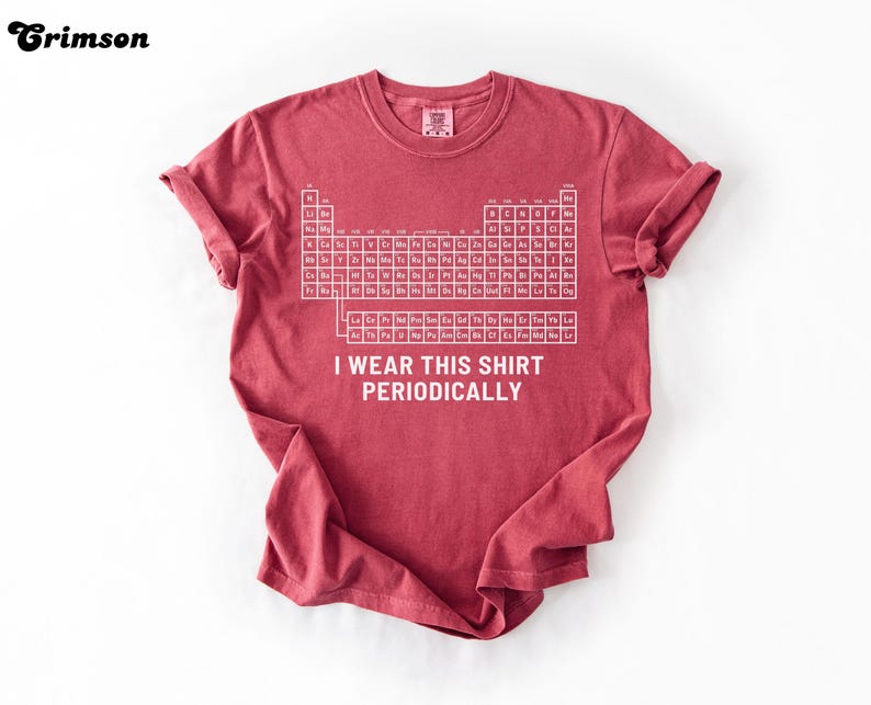 May include: Crimson-colored t-shirt featuring a white periodic table of elements graphic. The text "I wear this shirt periodically" is printed below the table. The shirt has rolled-up sleeves and a crew neck.