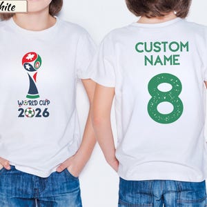 May include: A white short-sleeved t-shirt featuring a World Cup 2026 graphic on the front with a soccer ball and maple leaf icon. The back of the shirt displays the text CUSTOM NAME in green above a large green number 8 with a distressed texture.