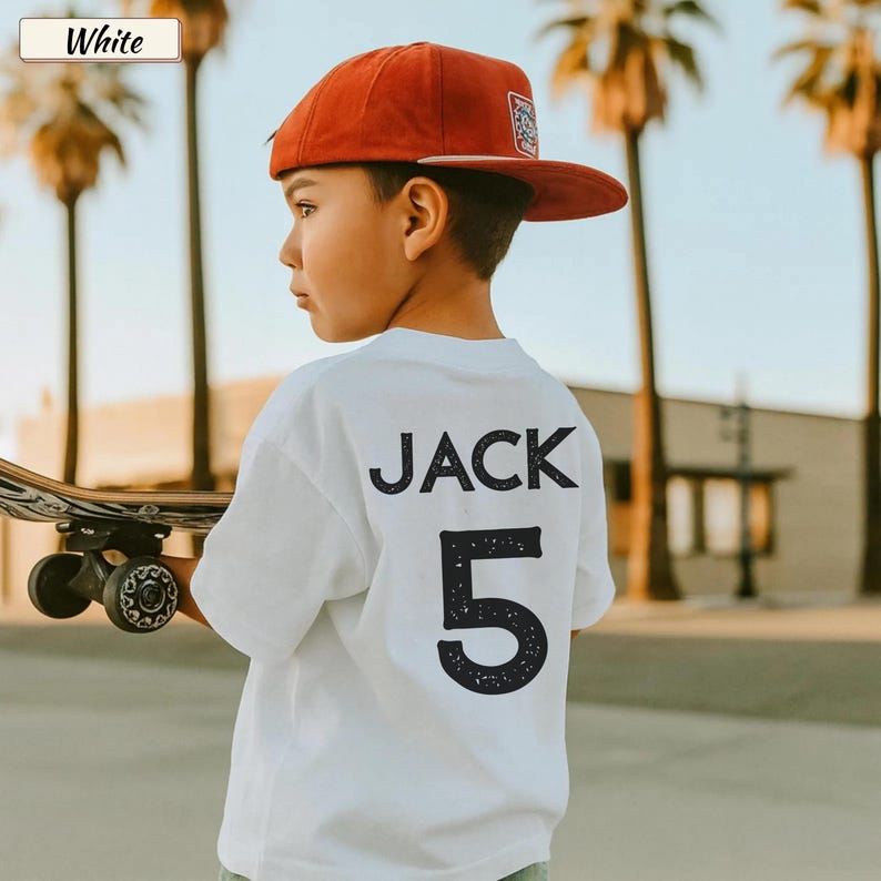 May include: A white short-sleeved t-shirt featuring the name JACK and the number 5 printed in a dark, distressed font on the back. The shirt is worn with a solid red baseball cap with a flat brim and a small patch on the front panel.