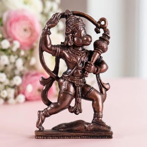 May include: A bronze-colored metallic figurine of the deity Hanuman in a dynamic standing pose, holding a mace over one shoulder. The figure features intricate detailing on the crown, jewelry, and clothing, set on a rectangular base.